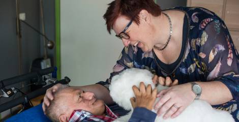 Man in bed met knuffel, beleidster heeft hand op zijn hoofd, ze lachen naar elkaar